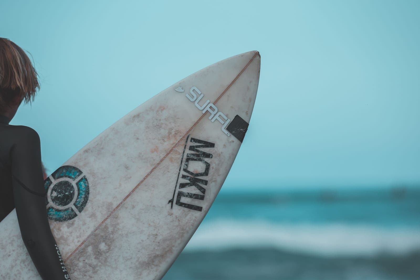 Eine Person im Neoprenanzug trägt ein Surfbrett Richtung Meer, im Hintergrund unscharfe Wellen