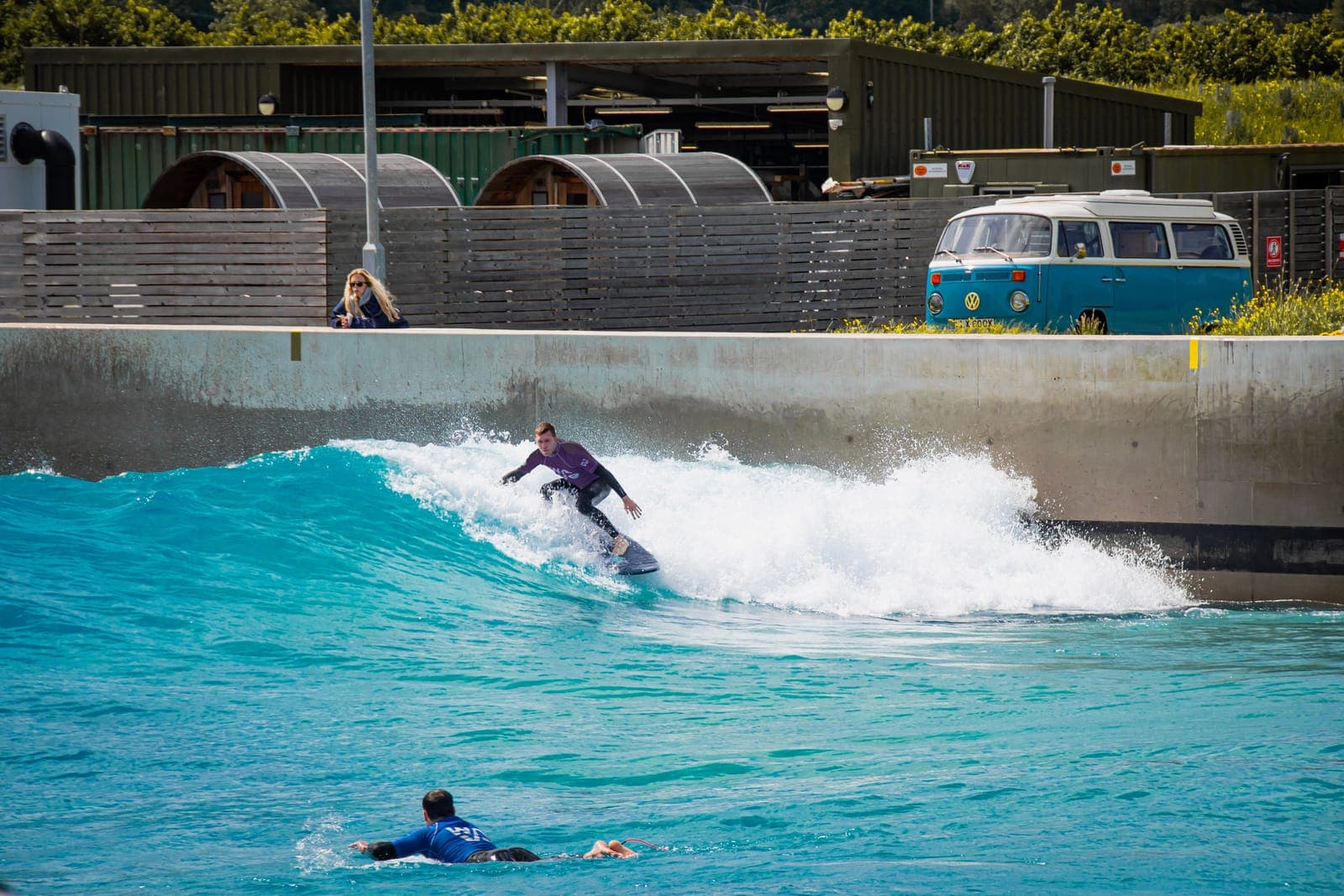 Ein Surfer fährt eine brechende Welle in einem Wellenbecken, im Hintergrund ein VW-Bulli und Holzkabinen am Beckenrand