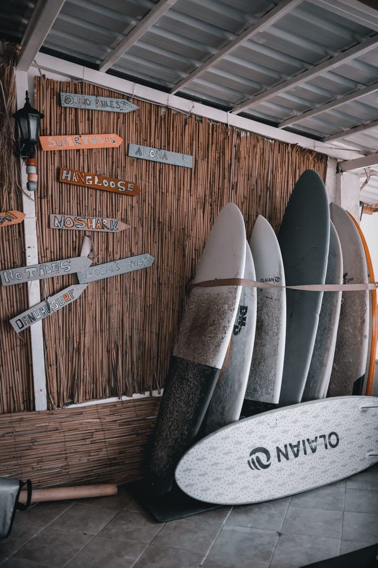 Reihe von Surfbrettern lehnt an einer Bambuswand mit handgemalten Schildern wie „Hangloose“ und „Aloha“